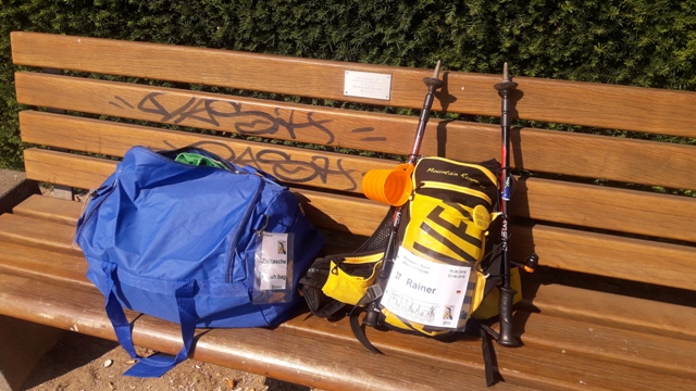 Auf der Bank liegen die gepackte Tasche für die beiden großen Versorgungspunkte (Dropbag) und der Laufrucksack. Außen angebracht die Stöcke und ein Trinkgefäß.
